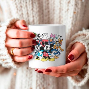 May include: White ceramic mug featuring a colorful illustration of Mickey Mouse, Minnie Mouse, Donald Duck, Daisy Duck, Goofy, and Pluto. The characters are depicted in their classic outfits. The mug is held by a person.