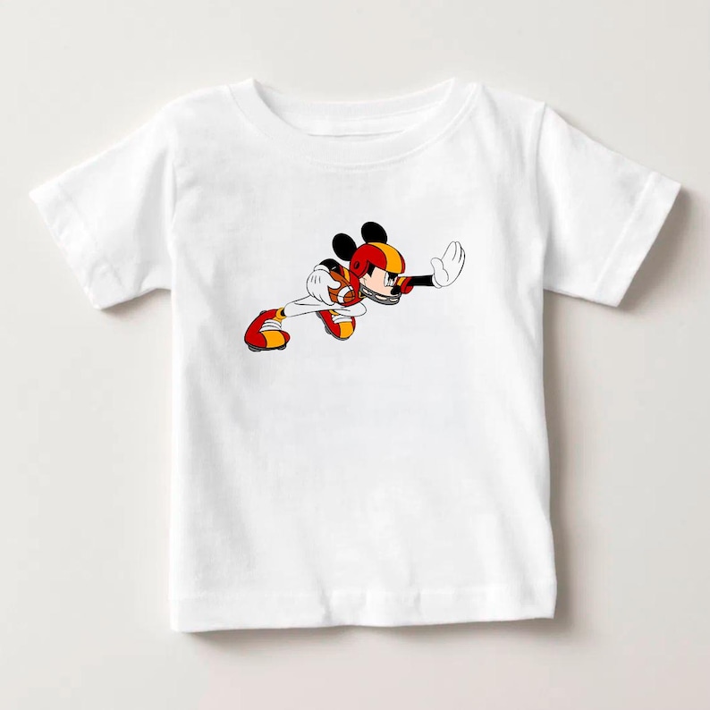 May include: White short-sleeved t-shirt featuring a cartoon illustration of Mickey Mouse in a football uniform. The character is depicted in a dynamic pose, wearing a red, yellow, and black helmet and uniform. The design is centered on the front of the shirt.