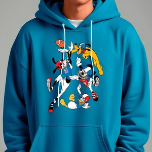 May include: Teal blue hoodie featuring a cartoon illustration of Mickey Mouse, Donald Duck, and Pluto playing football. The graphic includes footballs and helmets. The hoodie has a drawstring hood and a kangaroo pocket.
