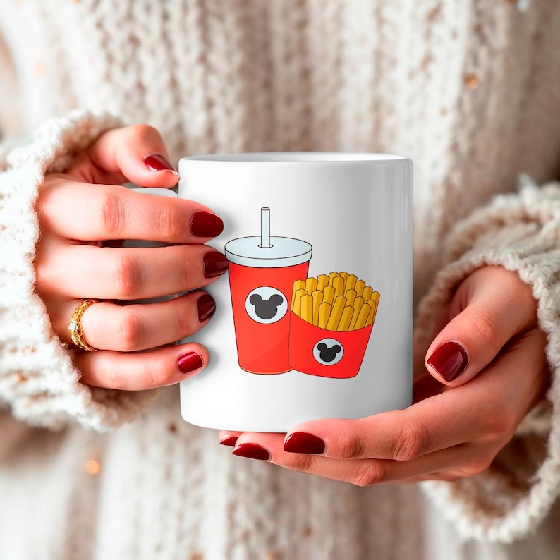 May include: White ceramic mug featuring a red soda cup and a red container of french fries, both with a black Mickey Mouse silhouette. The mug is held by a person wearing a cream-colored sweater.