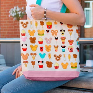 May include: A cream-colored tote bag with a pink bottom and teal straps. The bag is covered in colorful cartoon illustrations of various food items, including Mickey Mouse-shaped waffles, ice cream cones, and french fries. The bag is being held by a person.