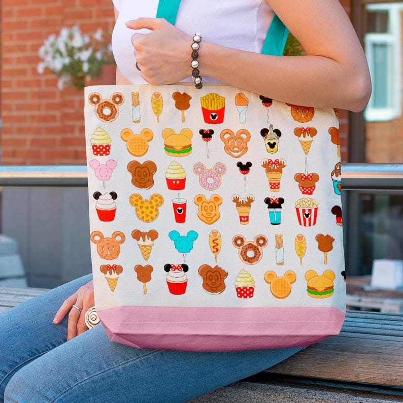 May include: A cream-colored tote bag with a pink bottom and teal straps. The bag is covered in colorful cartoon illustrations of various food items, including Mickey Mouse-shaped waffles, ice cream cones, and french fries. The bag is being held by a person.