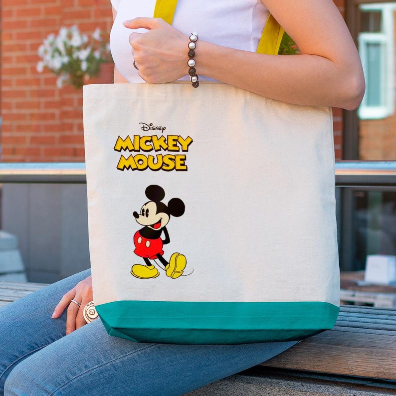 May include: Cream-colored tote bag featuring the Disney Mickey Mouse logo and a cartoon image of Mickey Mouse. The bag has a teal green bottom and yellow straps. The bag is made of a sturdy canvas material.
