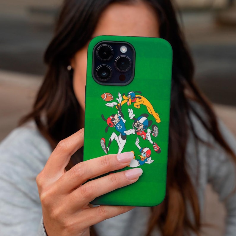 May include: Green phone case featuring a cartoon illustration of Mickey Mouse, Goofy, and Donald Duck playing football. The case has a green background with white lines, resembling a football field. The characters are in football uniforms.