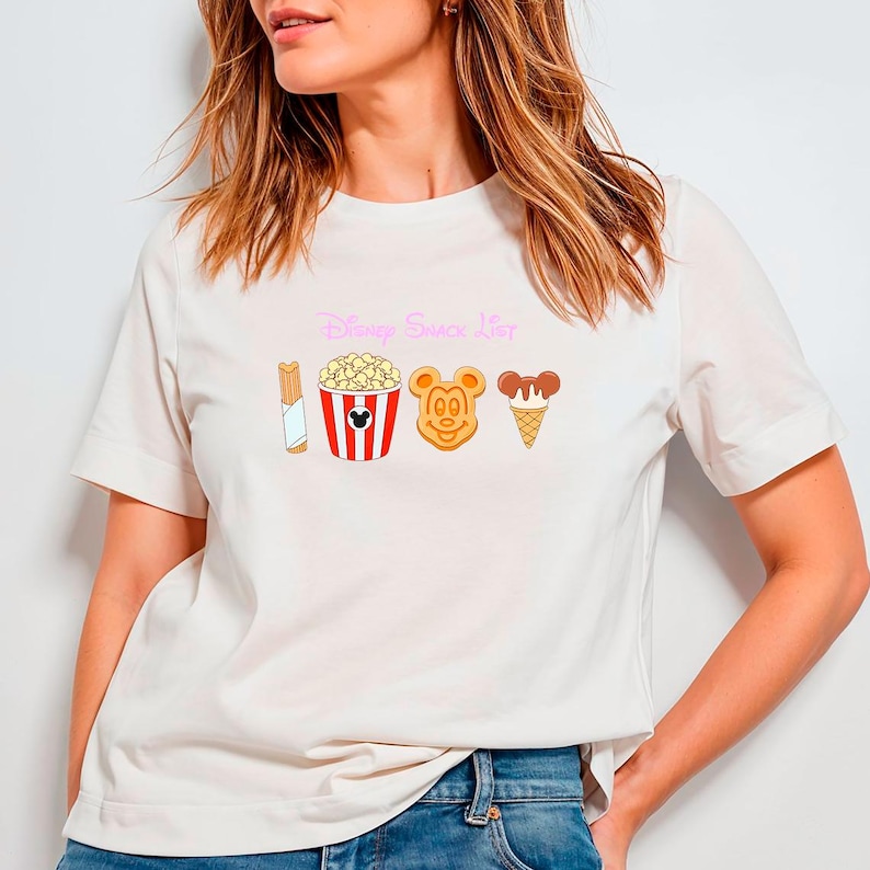 May include: Off-white t-shirt featuring the text "Disney Snack List" in pink script above cartoon illustrations of a churro, popcorn, Mickey Mouse-shaped waffle, and ice cream cone. The shirt has short sleeves.