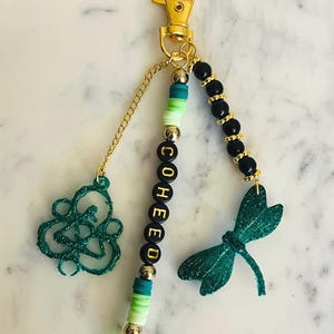 Può includere: Un portachiavi color oro con moschettone. Il portachiavi presenta un ciondolo a forma di libellula verde glitterata, un ciondolo astratto verde e una fila di perline con la scritta "COHED". Perline aggiuntive sono nere e dorate.