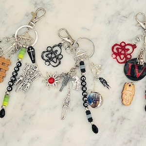 Puede incluir: Tres llaveros con varios dijes, incluyendo una libélula, un esqueleto en una motocicleta y una guillotina. Cada llavero tiene el nombre "COHEED" escrito con cuentas. Los dijes son plateados, negros, rojos y verdes.