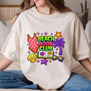 Peut inclure: T-shirt crème avec un motif coloré "Beach Book Club". Le design comprend un maillot de bain rose à pois, un coquillage, une étoile de mer, des lunettes de soleil, un palmier et un livre ouvert. Le texte est en vert, violet et jaune.
