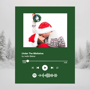 Könnte beinhalten: Ein weihnachtliches Bild mit einem küssenden Paar unter Mistelzweigen, das Weihnachtsmützen und Pullover trägt. Der Hintergrund ist ein verschneiter Wald. Der Text "Under The Mistletoe" von Justin Bieber wird zusammen mit einer Musikplayer-Oberfläche angezeigt.