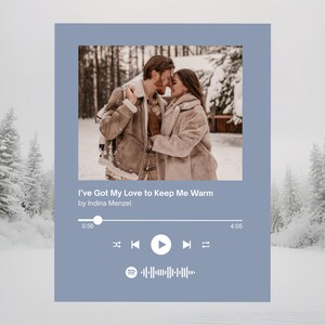 Könnte beinhalten: Digitaler Kunstdruck mit einem sich umarmenden Paar in einem verschneiten Wald. Das Bild ist in einer hellblauen Grafik im Spotify-Stil mit dem Text "I've Got My Love to Keep Me Warm" und Musiksteuerungen gerahmt. Der Hintergrund zeigt schneebedeckte Bäume.
