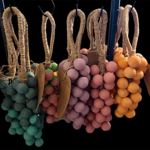 Op de afbeelding: Een verzameling decoratieve druiventrossen in verschillende kleuren, waaronder teal, rood, paars, roze en oranje. Elke tros is gemaakt van kleine, ronde bollen en heeft een lus van jute touw om op te hangen. De achtergrond is zwart.