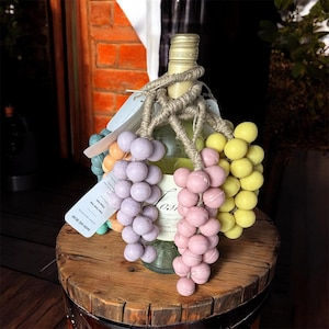 Puede incluir: Una botella de vino blanco adornada con racimos de uvas decorativas en tonos morado, rosa y amarillo. La botella está atada con hilo y descansa sobre un barril de madera. Una etiqueta está adjunta a la botella.