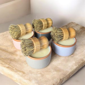 Può includere: Quattro piccoli portasapone rotondi in ceramica dai colori pastello, ciascuno con una saponetta e una spazzola per piatti in legno. I piatti hanno un bordo colorato chiaro e sono disposti su una superficie chiara e strutturata.
