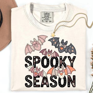 May include: A cream-colored t-shirt featuring a graphic print of four stylized bats with patterned wings and bows. The text "SPOOKY SEASON" is printed in large black letters decorated with small floral accents. The shirt has a Comfort Colors label.