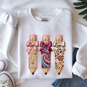 May include: A white crewneck sweatshirt featuring a graphic of three pencils, each topped with a satin bow. The first pencil has gold glitter, the second a colourful swirl pattern, and the third a leopard print with floral accents. The Gildan label is visible.
