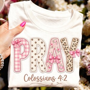 Könnte beinhalten: Ein weißes T-Shirt mit dem Wort PRAY in großen, stilisierten Buchstaben. Jeder Buchstabe hat ein einzigartiges Muster: rosa Vichy-Karo, braune Punkte, rosa Blumen und Leopardenmuster. Zwei rosa Schleifen sind angebracht, Colossians 4:2 steht darunter.