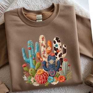 Peut inclure: Un sweat-shirt marron avec un motif de cactus coloré. Les cactus sont décorés de motifs floraux, de denim et d'un imprimé vache. Le motif comprend un fer à cheval et diverses fleurs.