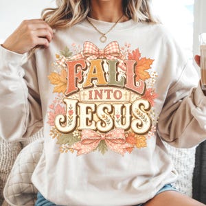 Op de afbeelding: Een beige sweater met ronde hals en een grafische print met de tekst "FALL INTO JESUS" in een gestileerd lettertype. Het ontwerp is omringd door herfstbladeren, kleine bloemen en twee roze gingham strikken, met kleine kruisjes in de tekst.