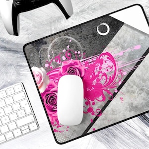May include: A rectangular mouse pad featuring a graphic design of vibrant pink roses and swirling floral patterns set against a gray concrete-textured background. A solid white computer mouse is positioned on the center of the mouse pad.