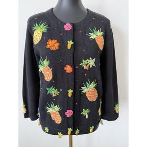 May include: Black cardigan sweater with embroidered pineapples, flowers, and palm trees. The sweater features a button-up front, long sleeves, and beaded trim. The pineapples are yellow and orange, and the flowers are pink and orange.