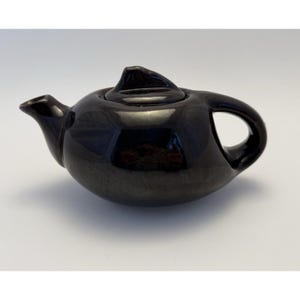 May include: A small, black ceramic teapot with a rounded body, a curved spout, and a loop handle. The lid has a pointed finial. The teapot has a glossy finish and reflects the surrounding light. The teapot is likely for brewing tea.