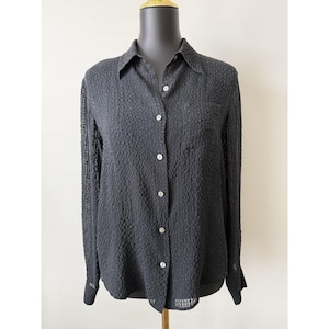 May include: A black long-sleeved button-down shirt featuring a textured, puckered fabric surface. The garment includes a standard collar, a single chest pocket on the left side, and light-colored buttons running down the center front placket.