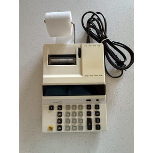 Puede incluir: Calculadora vintage de color crema con un soporte para rollo de papel. La calculadora tiene teclas negras con números y símbolos, y un cable de alimentación negro. El modelo "110" está impreso en la calculadora.