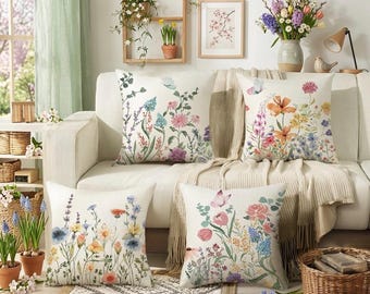 May include: A set of four square throw pillows featuring off-white fabric covers printed with colorful watercolor-style illustrations of various wildflowers, green foliage, and small butterflies in shades of pink, blue, orange, and yellow.