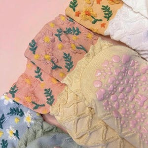 May include: Close-up of several pairs of socks in various colors, including pink, yellow, and white. The socks feature floral and leaf patterns, with some having textured soles. The socks are made of a soft material.