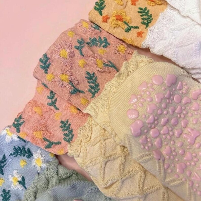 May include: Close-up of several pairs of socks in various colors, including pink, yellow, and white. The socks feature floral and leaf patterns, with some having textured soles. The socks are made of a soft material.