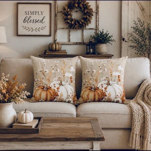 May include: Two square throw pillows featuring a printed design of orange and white pumpkins surrounded by dried wheat, cotton stems, and autumn foliage on a light beige background, displayed on a neutral-colored sofa with a textured throw blanket.