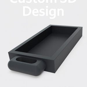 May include: A dark gray rectangular tray with a rounded handle on one end. The tray has a recessed interior and the text "Custom 3D Design" in white above it. The design is modern and minimalist.