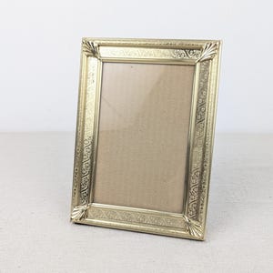 May include: A rectangular gold-toned metal picture frame featuring an intricate embossed floral pattern along the border and decorative fan-shaped accents at each of the four corners. The frame includes a clear glass pane and a plain brown backing.