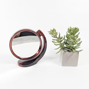 May include: A round, tortoiseshell-patterned vanity mirror with a pivoting frame. The mirror is positioned next to a small, square concrete planter holding a green succulent plant. The background is plain white.