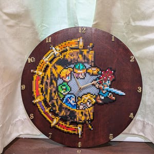 May include: A circular dark brown wooden wall clock featuring a pixel art design made of colorful plastic beads. The center displays a group of small pixelated characters in a circular arrangement, with gold-colored numbers marking the hours.