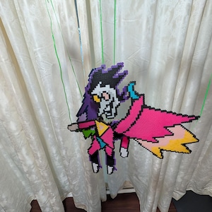 Puede incluir: Una figura de pixel art de un personaje con una cabeza negra y morada, una cara blanca y una capa rosa. La figura está hecha de cuentas de plástico de colores y está suspendida por una cuerda verde.