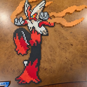 Può includere: Una figura pixel art di Blaziken, un Pokémon di tipo Fuoco, realizzata con perline di plastica colorate. La figura presenta una combinazione di colori rosso, nero, bianco e arancione, con fiamme che si estendono dalle sue braccia. La figura è esposta su una superficie di legno.