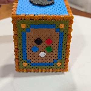 May include: A handmade cube-shaped decorative item made from colorful plastic beads. The top is blue with a black design, and the sides feature a brown background with a controller design. The edges are bordered with orange beads.