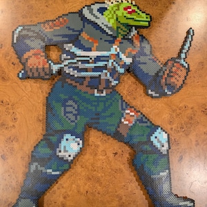 Può includere: Una figura in pixel art realizzata con perline di plastica fuse che raffigura una creatura verde simile a una lucertola con occhi rossi. Il personaggio indossa una giacca blu scuro con cappuccio, pantaloni mimetici e tiene in mano un oggetto appuntito azzurro.