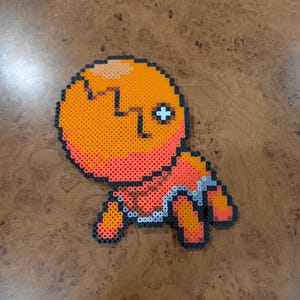 Puede incluir: Una figura de pixel art hecha de cuentas naranjas, rojas y negras. La figura tiene una cabeza redonda con un ojo en forma de cruz y un cuerpo con brazos y piernas pequeños. La figura está sobre una superficie de madera.