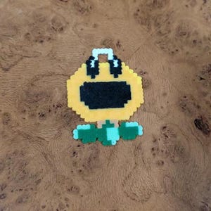 Op de afbeelding: Een gele pixel art zonnebloem met een zwarte mond en koptelefoon. De zonnebloem heeft groene bladeren en een lichtblauwe stengel. Het ontwerp is gemaakt van kleine plastic kralen.