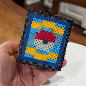 May include: A handmade, square-shaped bead art piece featuring a red and white ball design with a black center, surrounded by blue, yellow, and black beads. The artwork is made with small, colorful plastic beads.