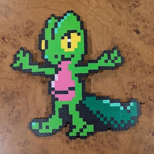 Pode incluir: Uma figura de pixel art feita de contas de plástico fundidas representando uma criatura verde semelhante a um lagarto com barriga rosa, olhos amarelos e cauda verde escura. O personagem está em pé com os braços estendidos e contornos pretos.