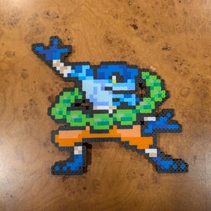 Może przedstawiać: Figura w stylu pixel art przedstawiająca niebiesko-zielone stworzenie z pomarańczowym pasem i czarnym konturem. Figurka wykonana jest z małych, kolorowych plastikowych koralików. Stworzenie jest w dynamicznej pozie, z uniesioną ręką.
