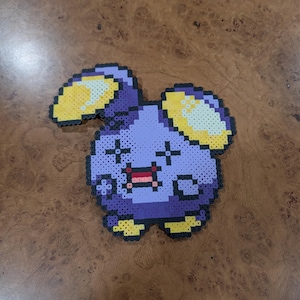May include: A pixel art character in purple, yellow, and black. The character has large ears, a small mouth, and crossed-out eyes, giving it a distressed expression. The piece is made of small, colourful beads, and is likely a handmade item.