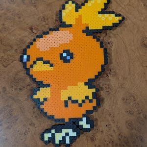 May include: A pixel art figure crafted from orange, yellow, and light yellow plastic beads, outlined in black. The figure depicts a stylized bird-like creature with a yellow crest, a rounded orange body, a yellow beak, and small yellow wings.