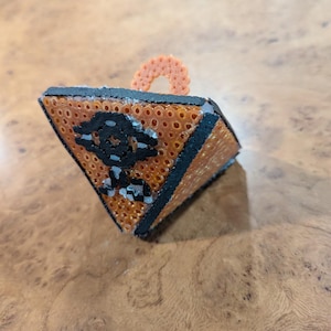 Op de afbeelding: Een geometrisch, driehoekig ornament met een zwart en oranje ontwerp. Het ornament heeft een kleine oranje lus aan de bovenkant. Het ontwerp heeft een zwart embleem aan de voorkant. Het ornament is gemaakt van kleine, ronde kralen.