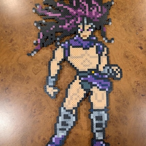 Puede incluir: Una figura de arte de píxeles de un personaje musculoso con cabello púrpura puntiagudo, vistiendo un atuendo púrpura y gris. La figura está hecha de pequeñas cuentas de colores y está posada en una postura dinámica. La figura está sobre una superficie de madera marrón.