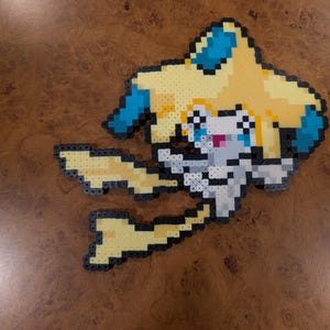 May include: A pixel art figure crafted from small plastic beads, depicting a stylized creature with a pale yellow star-shaped head, blue accents, and a white body. The character features a small pink mouth and dark outlines, creating a flat, grid-like appearance typical of fuse bead projects.