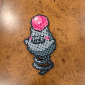 May include: A pixel art figure crafted from small plastic beads, depicting a grey creature with a rounded body, a pink circular pom-pom on its head, small pink cheek accents, and a coiled spring-like tail extending from the bottom of the figure.